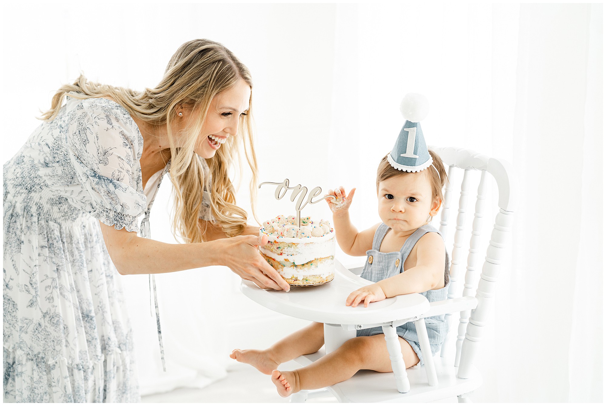 A mother present a first birthday cake to her baby seated in a high chair with a blue party hat on.
