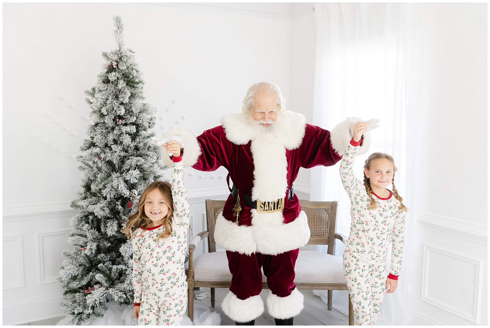 Two young girls wearing Christmas pajamas spin with Santa during an Atlanta Santa Photo session in a white studio.