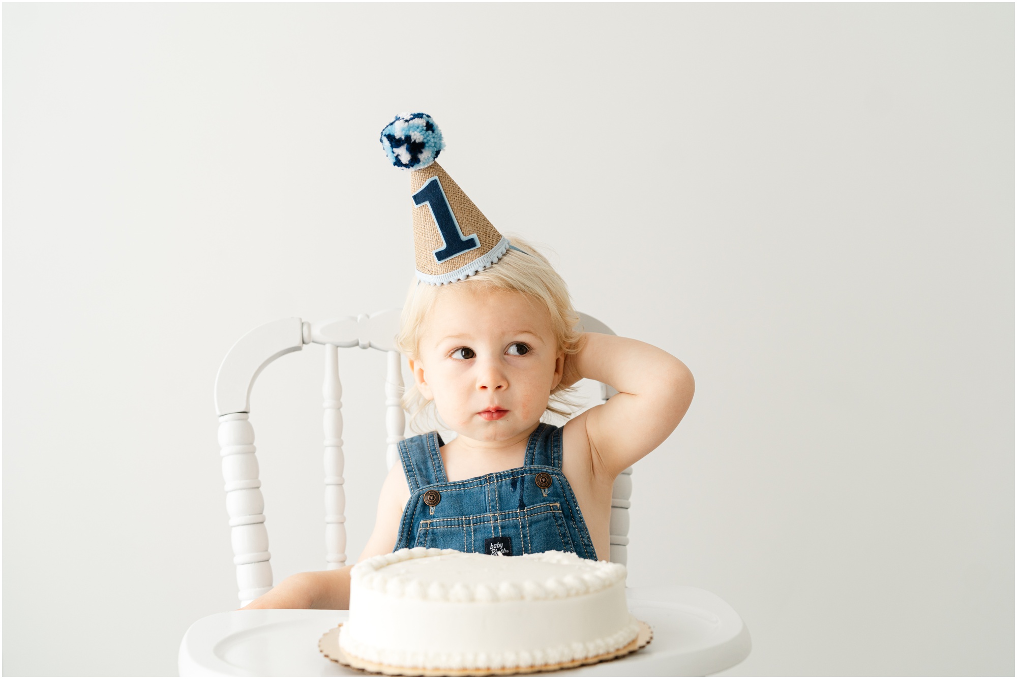 A young boy sits in a high chair with a cake in front of him and a birthday hate with a number one on it for a first birthday photo session.