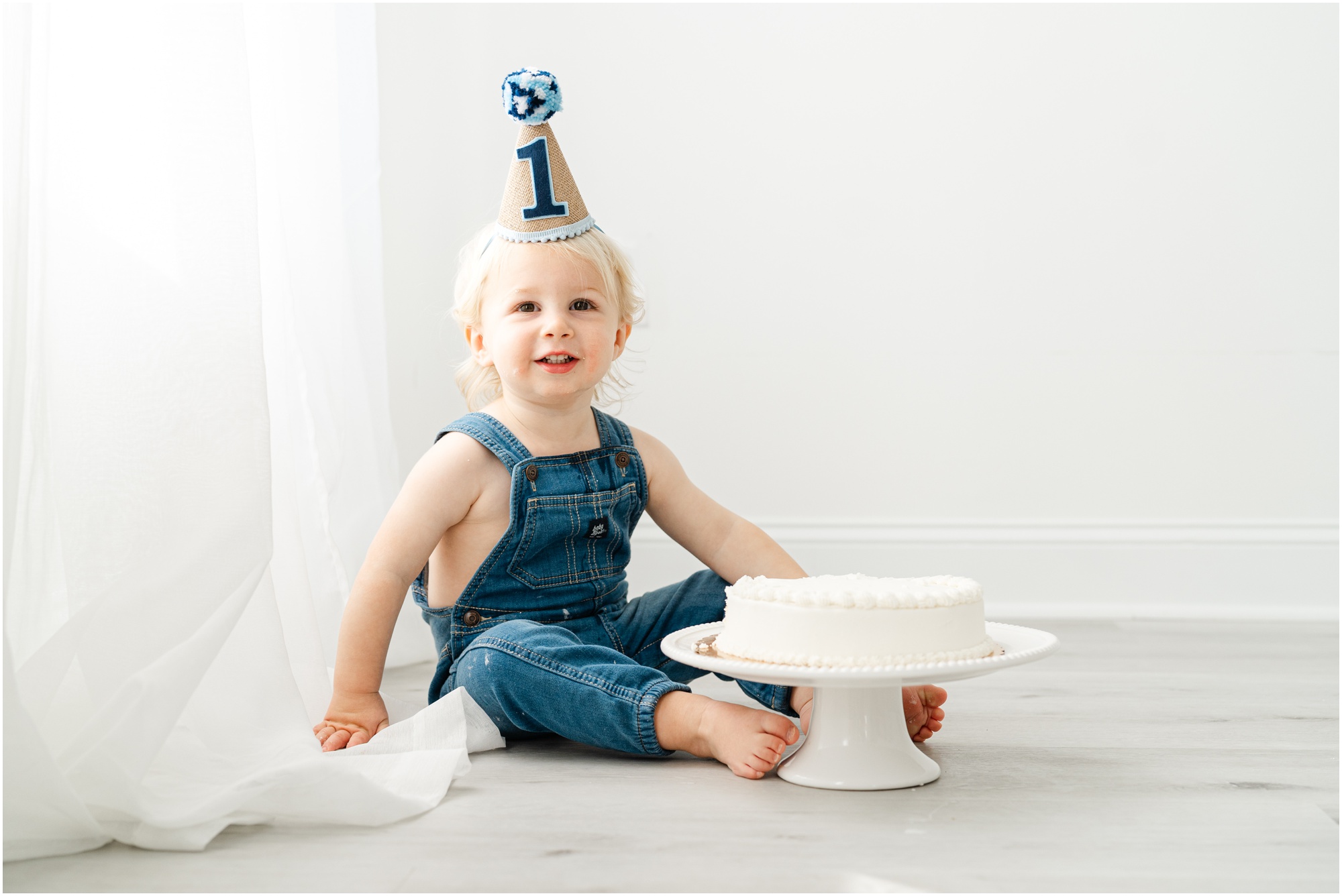 A young boy sits on the floor with a cake and a birthday hat on his head for his first birthday photo session.